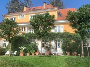 Ein zweistöckiges Gebäude mit rotem Ziegeldach, einem Balkon und einem Garten mit Kübelpflanzen und Gartenmöbeln.