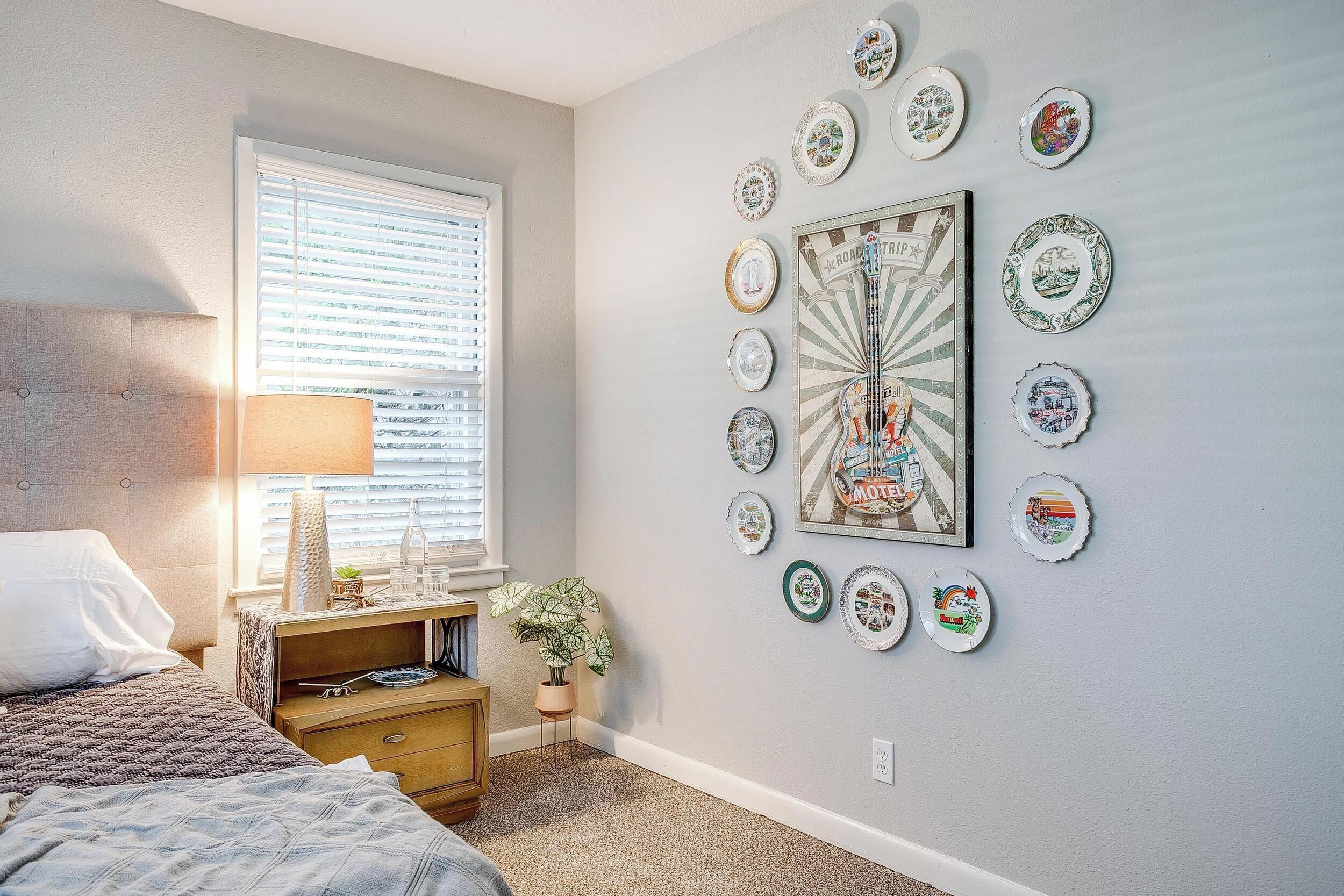 A bedroom with a bed, bedside table, lamp, and a wall decorated with plates and a vintage poster.