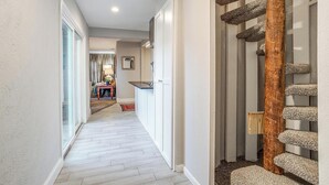 A hallway with a wooden floor, a staircase with a wooden handrail, and a kitchen area with a counter and cabinets. - The Westside Collective (Westworth Village)