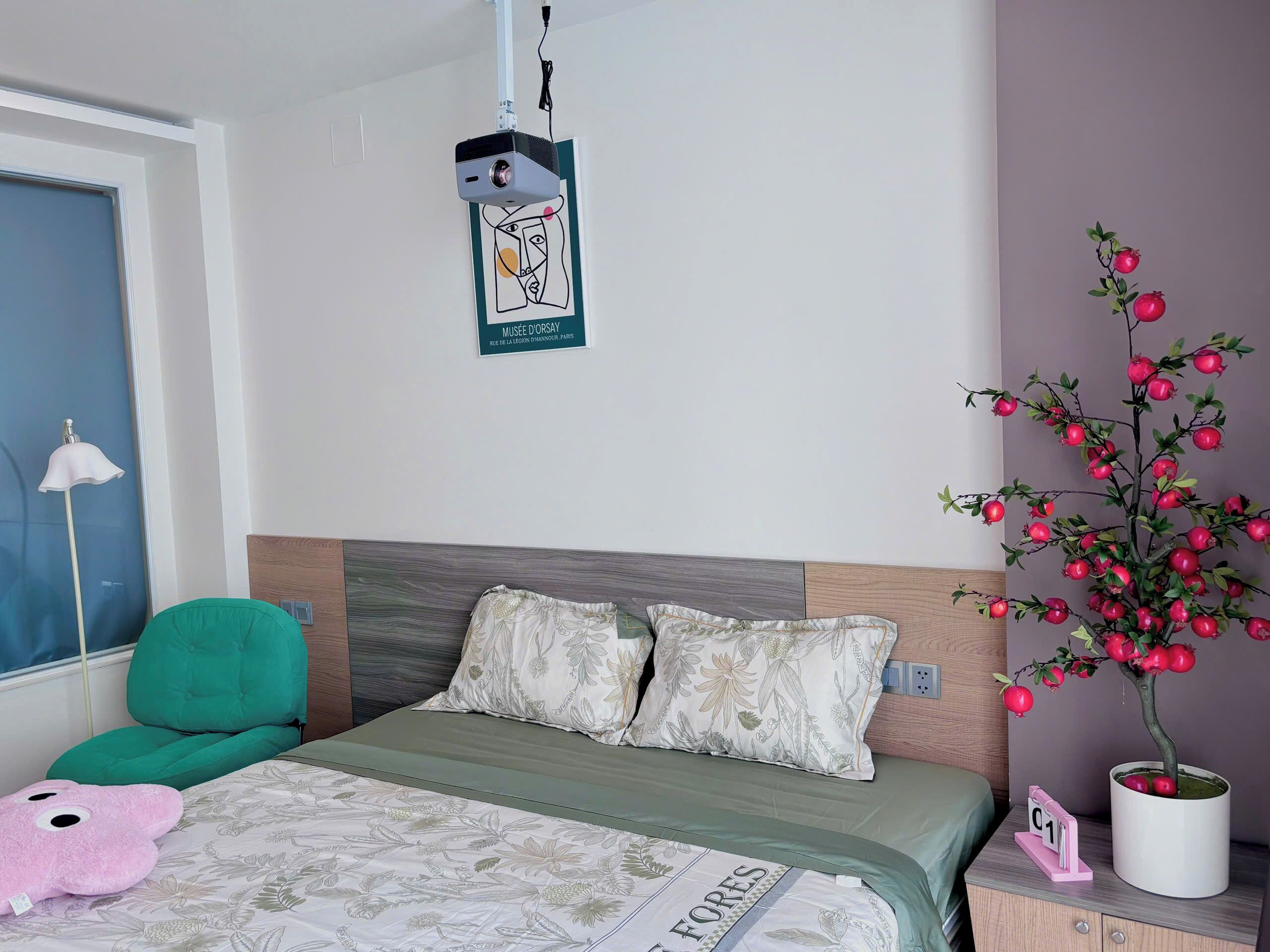 A neatly arranged bedroom with a bed, pillows, a green chair, a lamp, and a decorative pomegranate tree.
