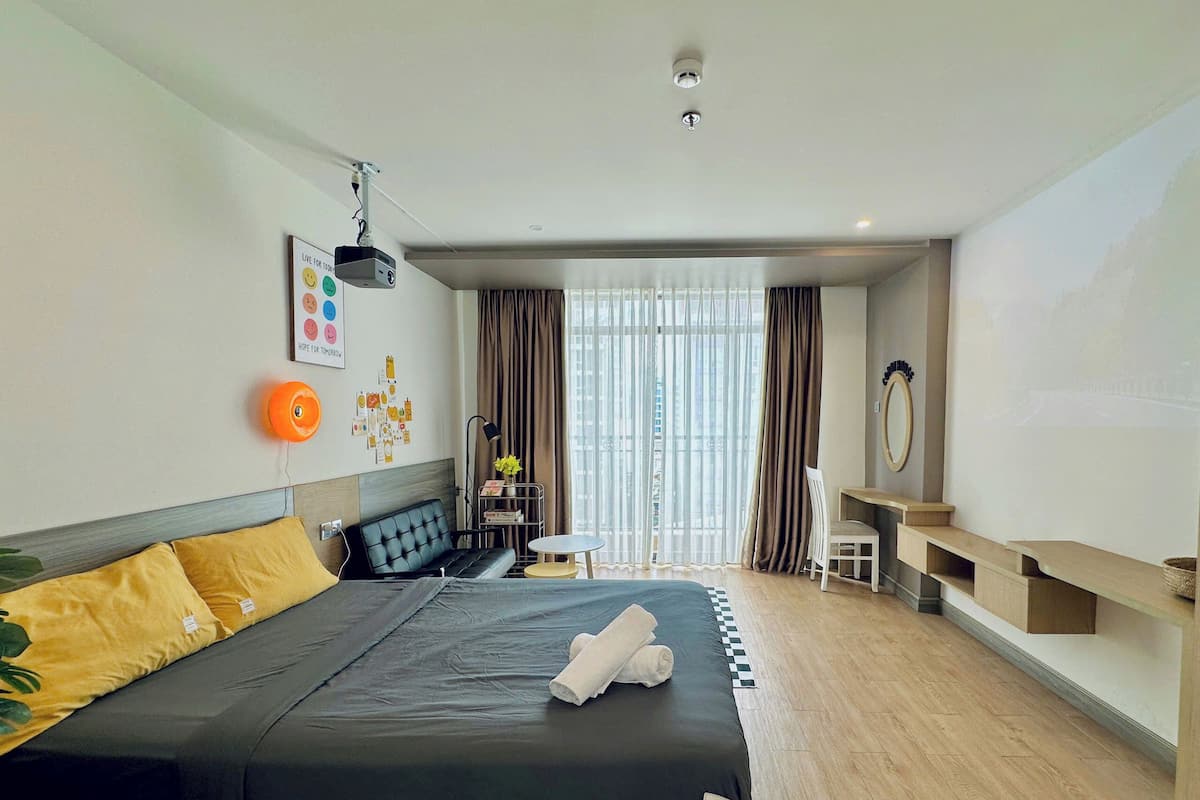 Ein Hotelzimmer mit einem großen Bett, einem Schreibtisch, einem Stuhl, einem kleinen Tisch, einem Spiegel und einem Fenster mit Vorhängen.