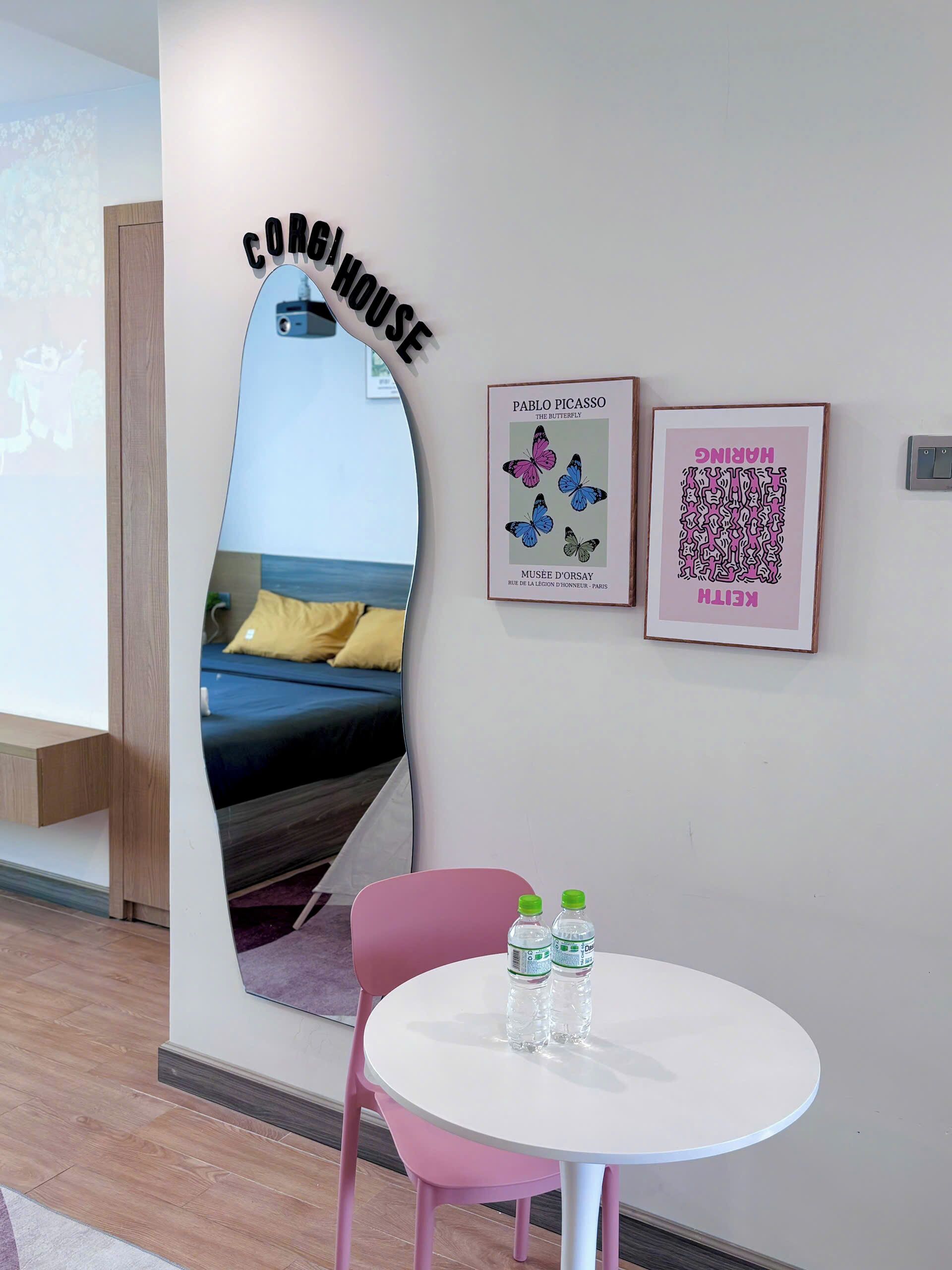 A modern room with a bed, a round table with water bottles, a pink chair, and two framed artworks on the wall.