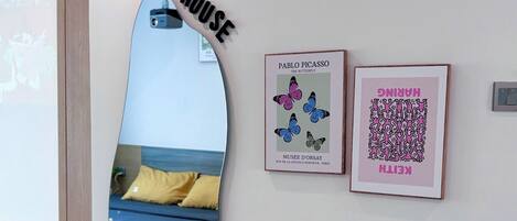 A modern room with a bed, a round table with water bottles, a pink chair, and two framed artworks on the wall.
