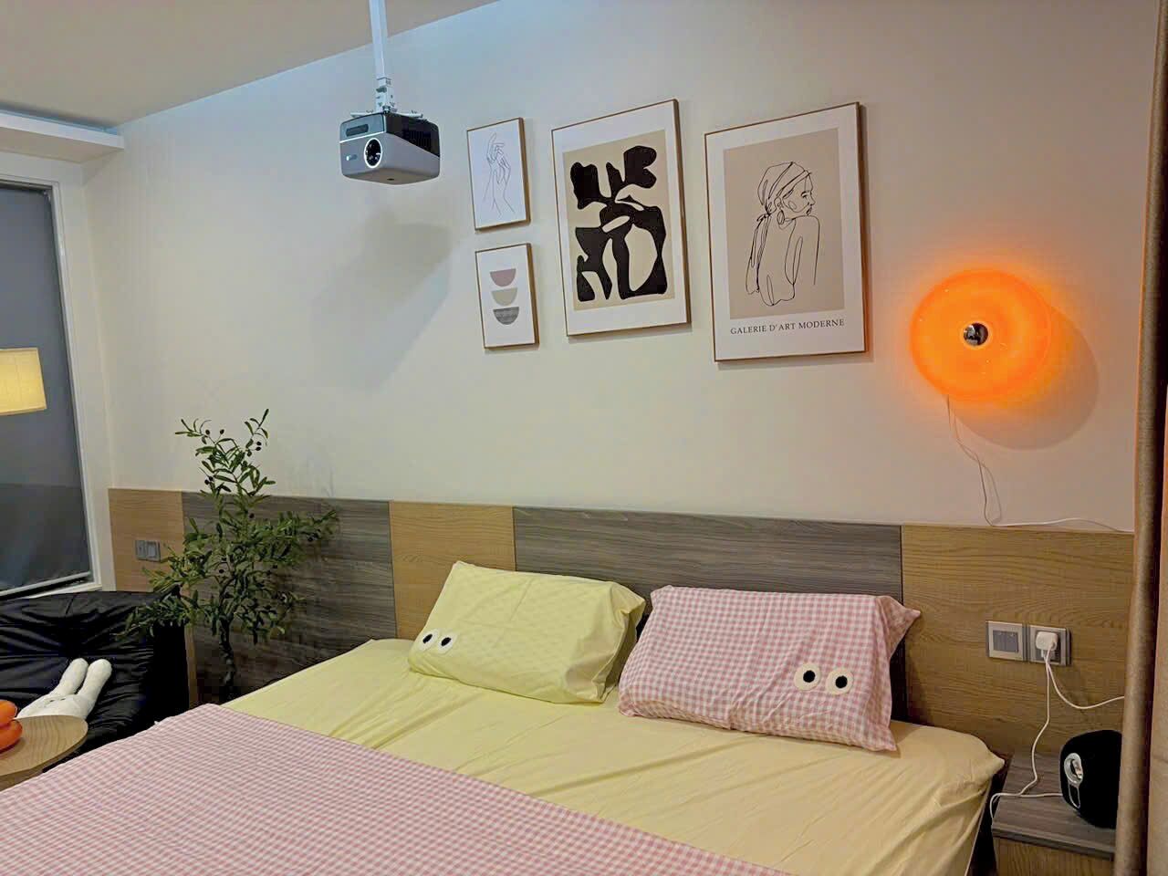 A hotel room with a bed, a projector, framed artwork, and a wall-mounted lamp.