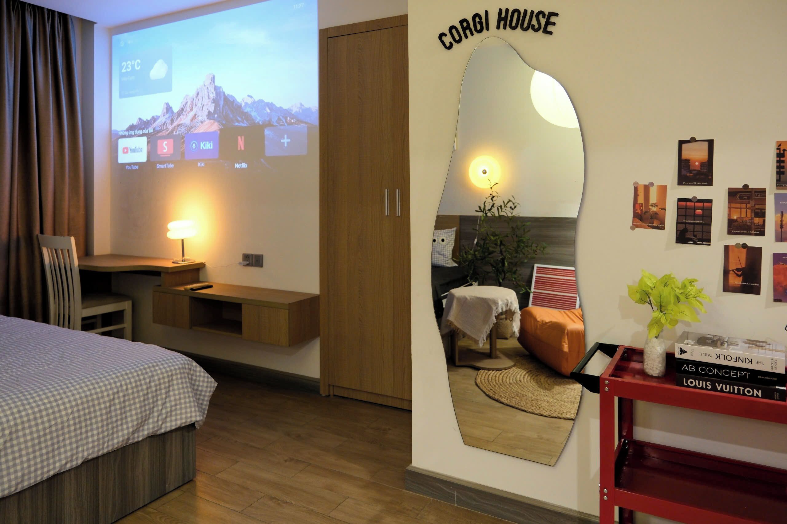 A hotel room with a bed, a desk, a TV screen displaying a mountain view, a mirror, and a wall with framed pictures.