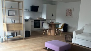 Un saló modern amb un sofà blanc, un otoman lila, un paviment de fusta, i una zona de cuina amb una taula de menjador i cadires.