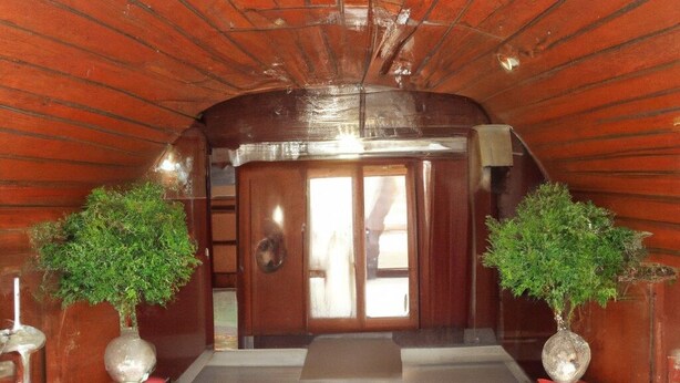 An arched wooden hallway with a tiled floor, two potted plants on red pedestals, and a door with a glass panel. - Giggler (Jamnagar)