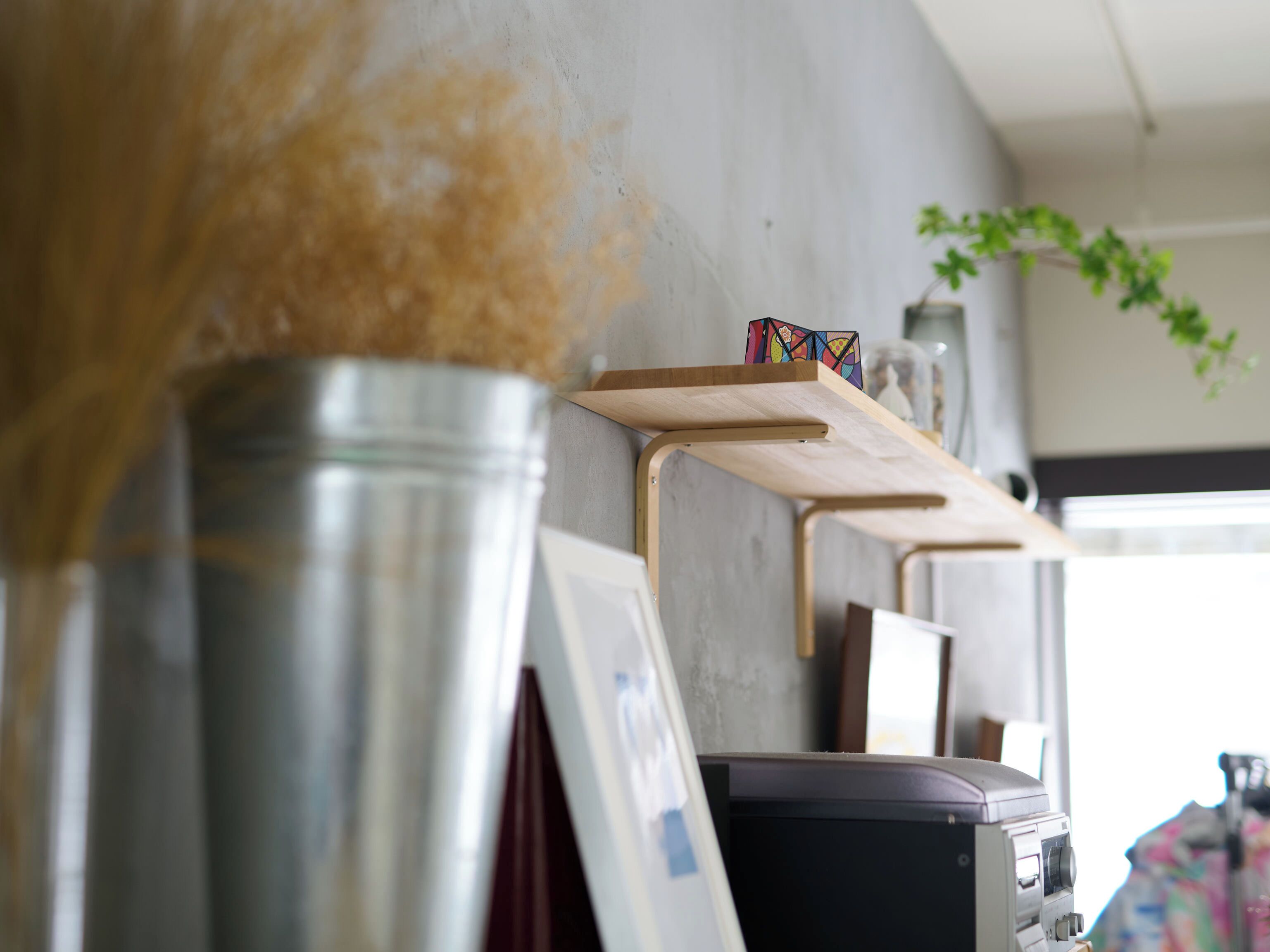 A metal bucket, a wooden shelf with a colorful object, and a plant on a wall.