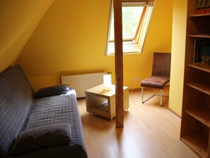 Una habitación pequeña y acogedora con techo inclinado, una cama individual, una silla, una mesa y una estantería.