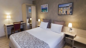 A hotel room with a bed, a desk, a chair, a bedside table with a lamp, and two paintings on the wall.