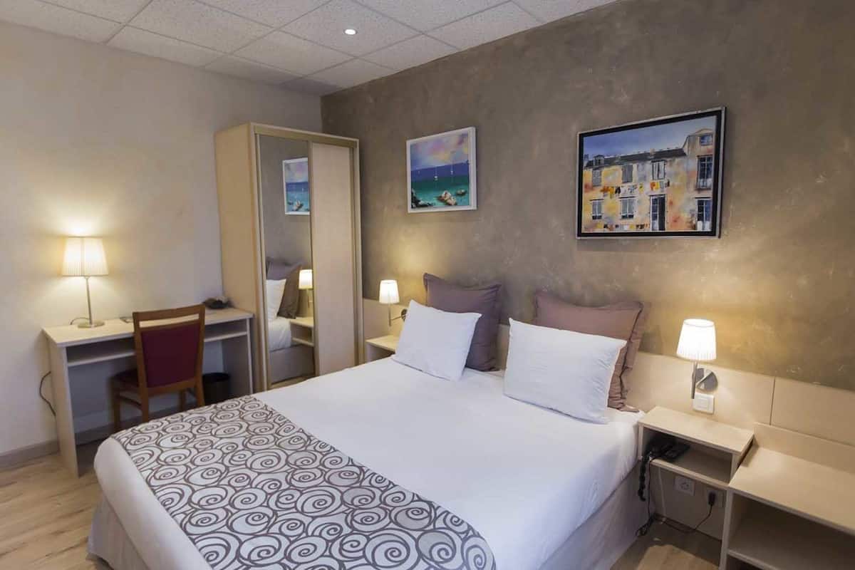A hotel room with a bed, a desk, a chair, a bedside table with a lamp, and two paintings on the wall.
