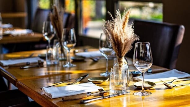 A restaurant interior with a long wooden table, chairs, and a vase with dried plants.