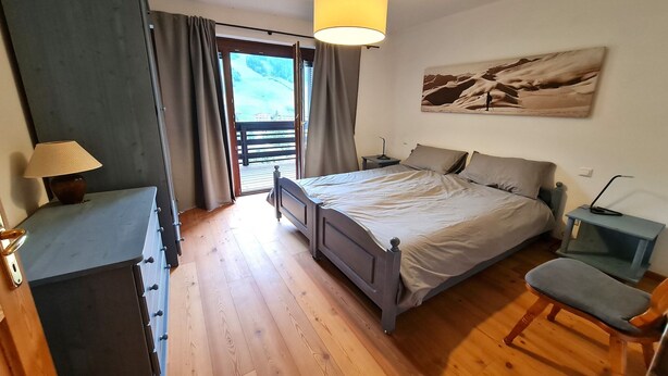 Ein Schlafzimmer mit Bett, Stuhl, Schreibtisch und Lampe. Es gibt ein Fenster mit Vorhängen und ein Bild an der Wand.