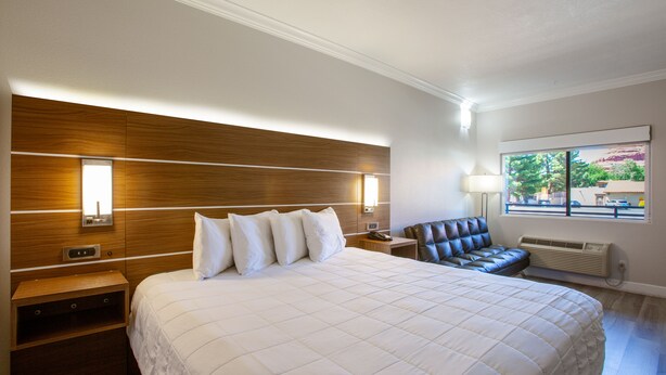 Quarto de hotel com uma cama grande, cabeceira de madeira, mesa de cabeceira, poltrona de couro e janela com vista para edifícios.