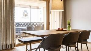 Un coin repas avec une table en bois, quatre chaises, un luminaire suspendu et une vue sur l’extérieur grâce à une fenêtre munie de stores.