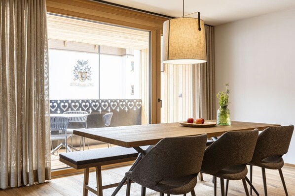 A dining area with a wooden table, four chairs, a hanging light fixture, and a view of the outside through a window with blinds.