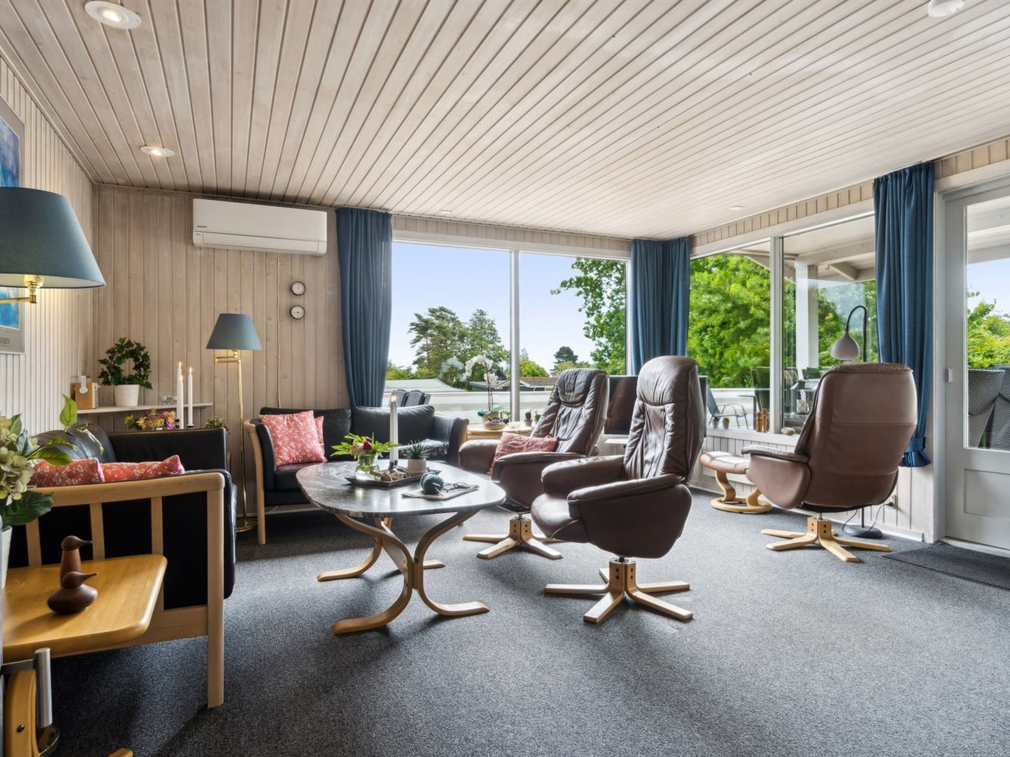 A spacious room with a wooden ceiling, large windows, and a view of greenery outside.