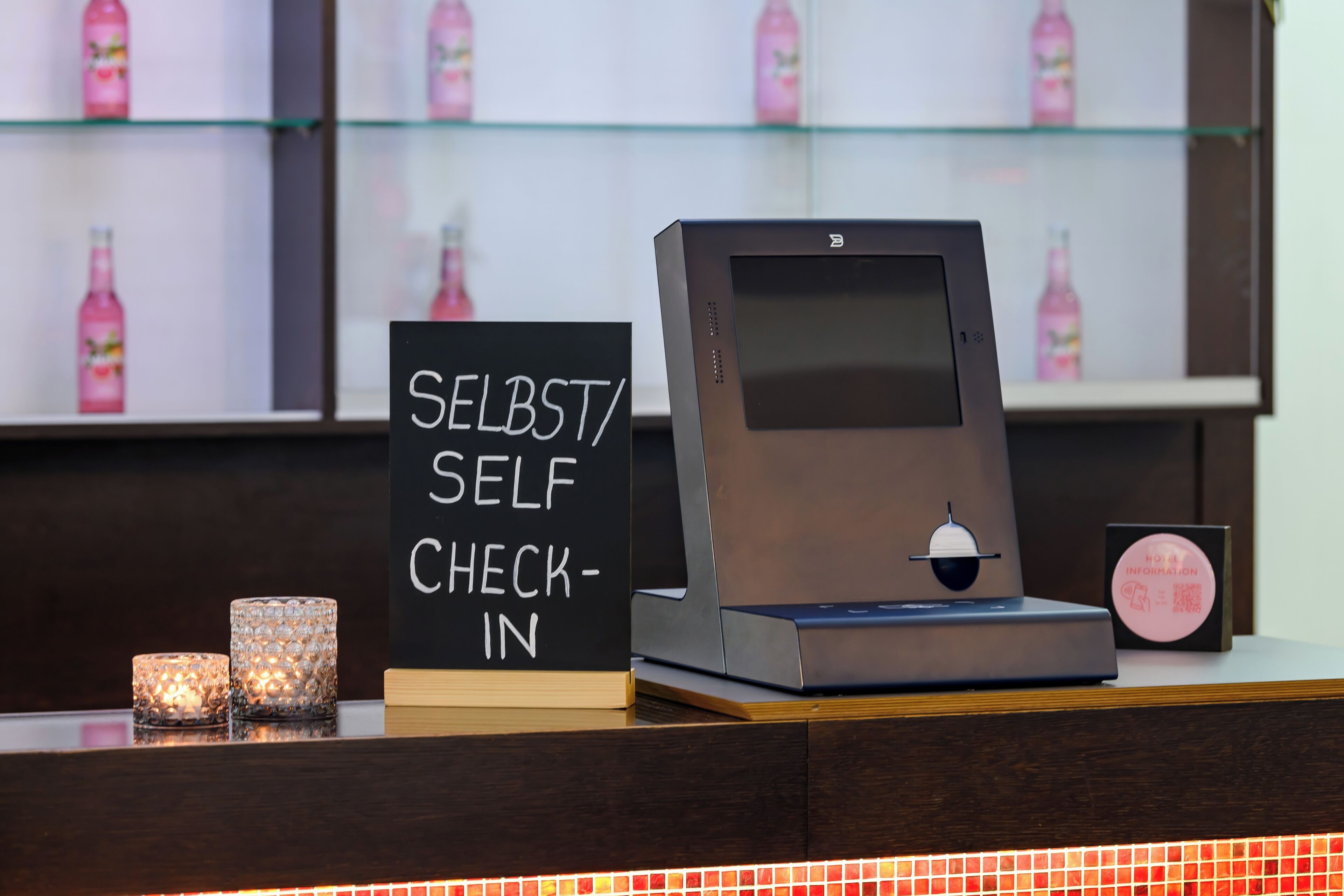 A self-check-in kiosk with a digital screen, a sign indicating self-check-in, and a display of bottled beverages in the background.