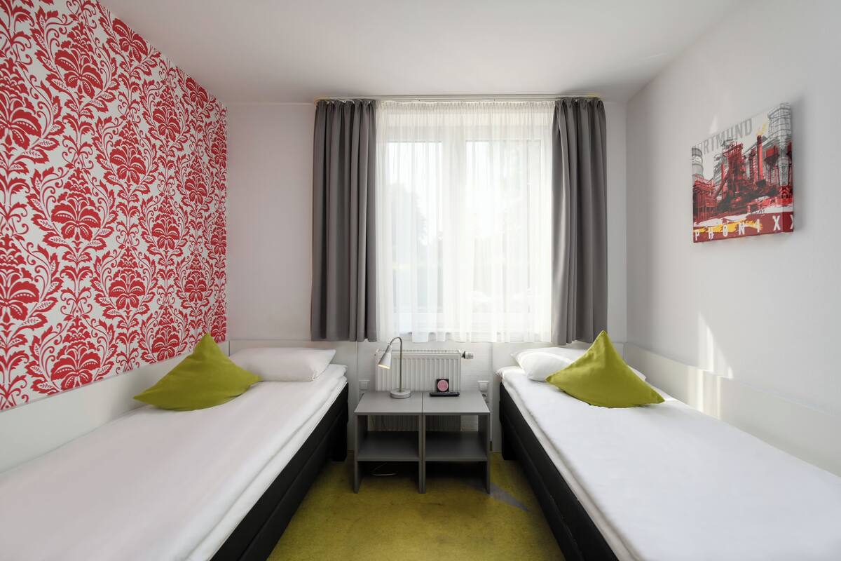 A room with two single beds, a small table with a lamp, a radiator, and a window with curtains.