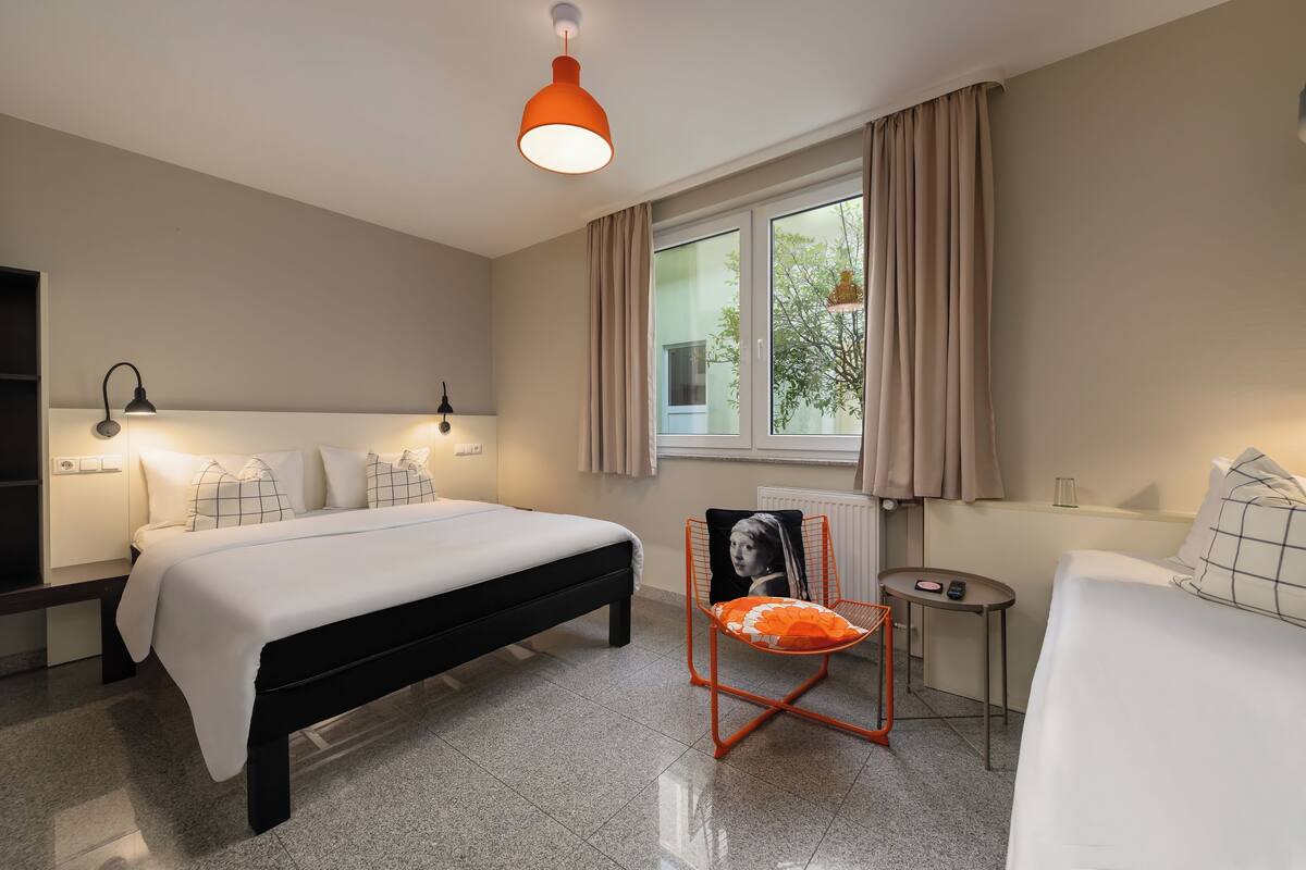 A modern hotel room with a large bed, a small desk, a chair with a cushion, and a window with curtains.