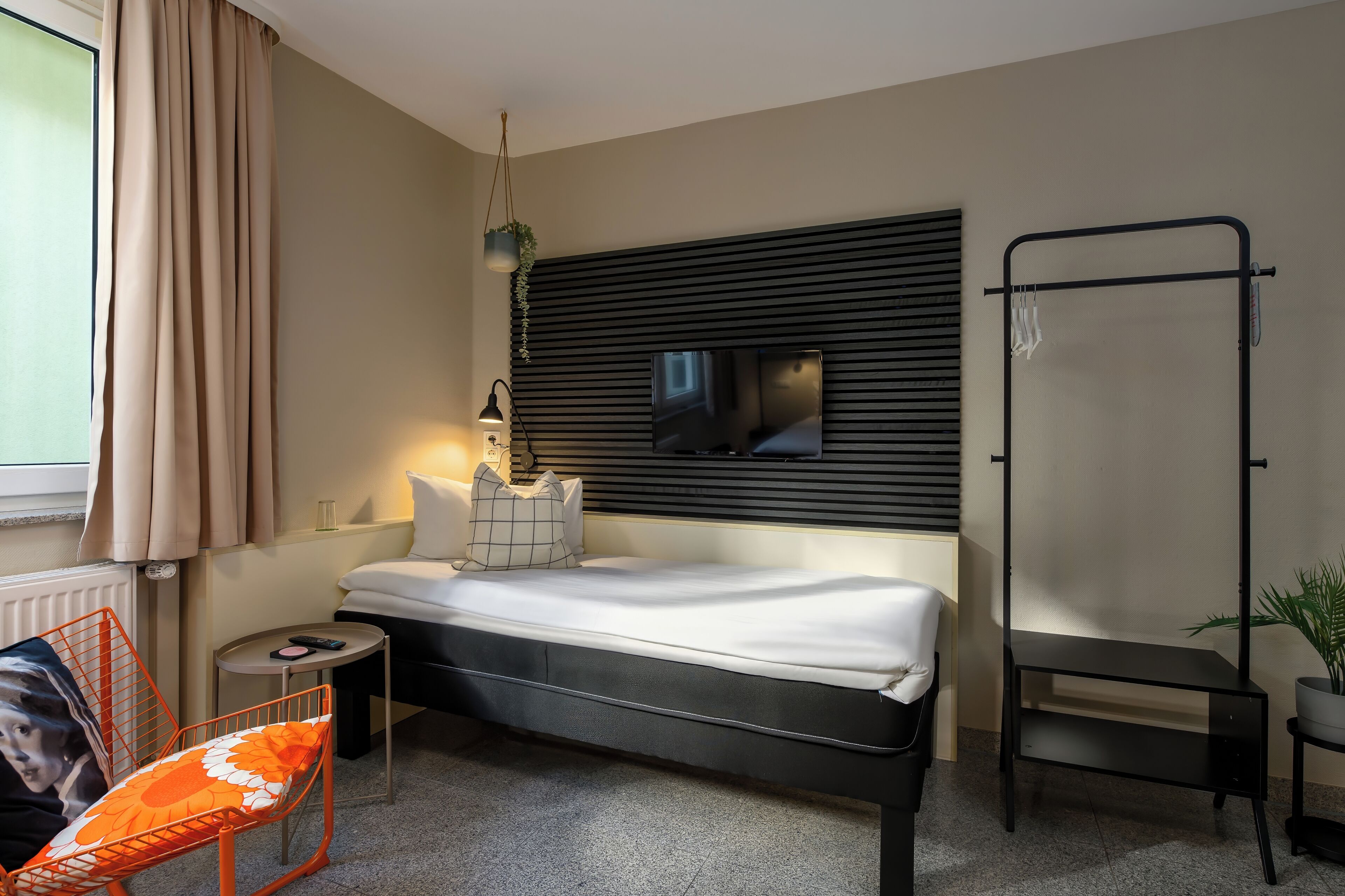 Een moderne hotelkamer met een bed, een televisie bevestigd aan een zwart paneel, een klein tafeltje met een lamp, een stoel met een bloemenkussen en een raam met gordijnen.