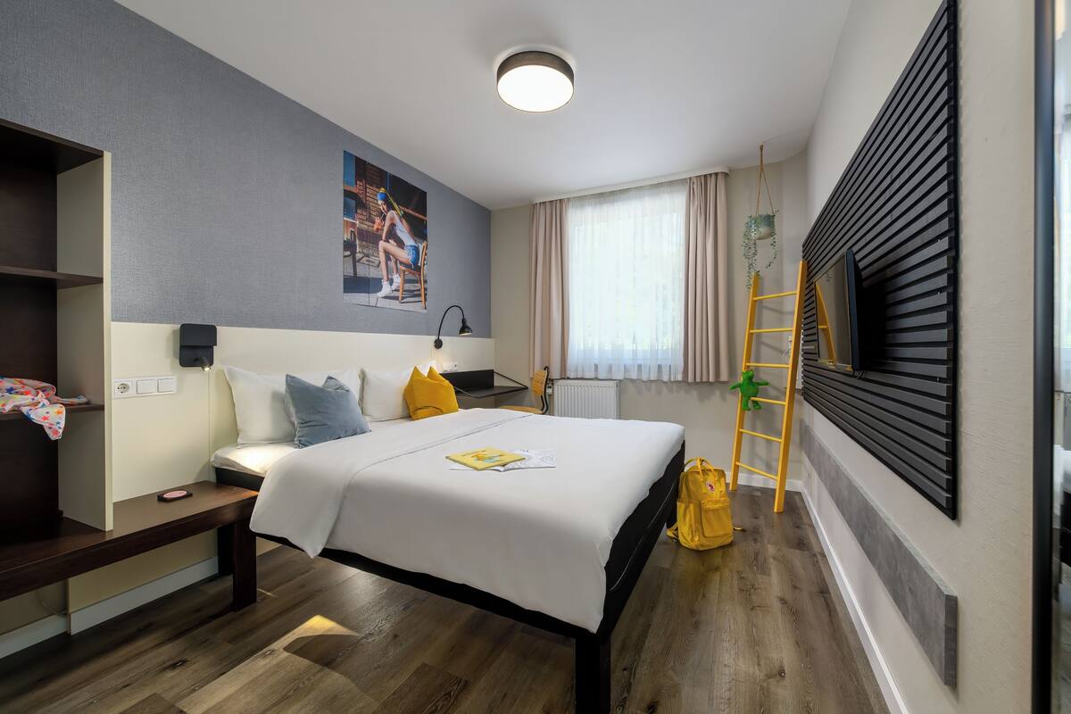 A modern hotel room with a bed, bedside table, lamp, and a view of the city through large windows.