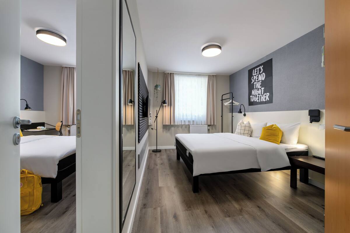 A modern hotel room with two beds, a desk, a chair, a mirror, a window with curtains, and a motivational wall quote.