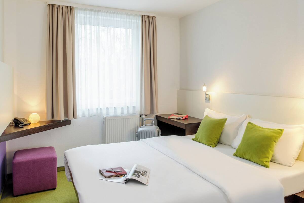 A hotel room with a bed, a bedside table, a lamp, a telephone, a radiator, and a window with curtains.