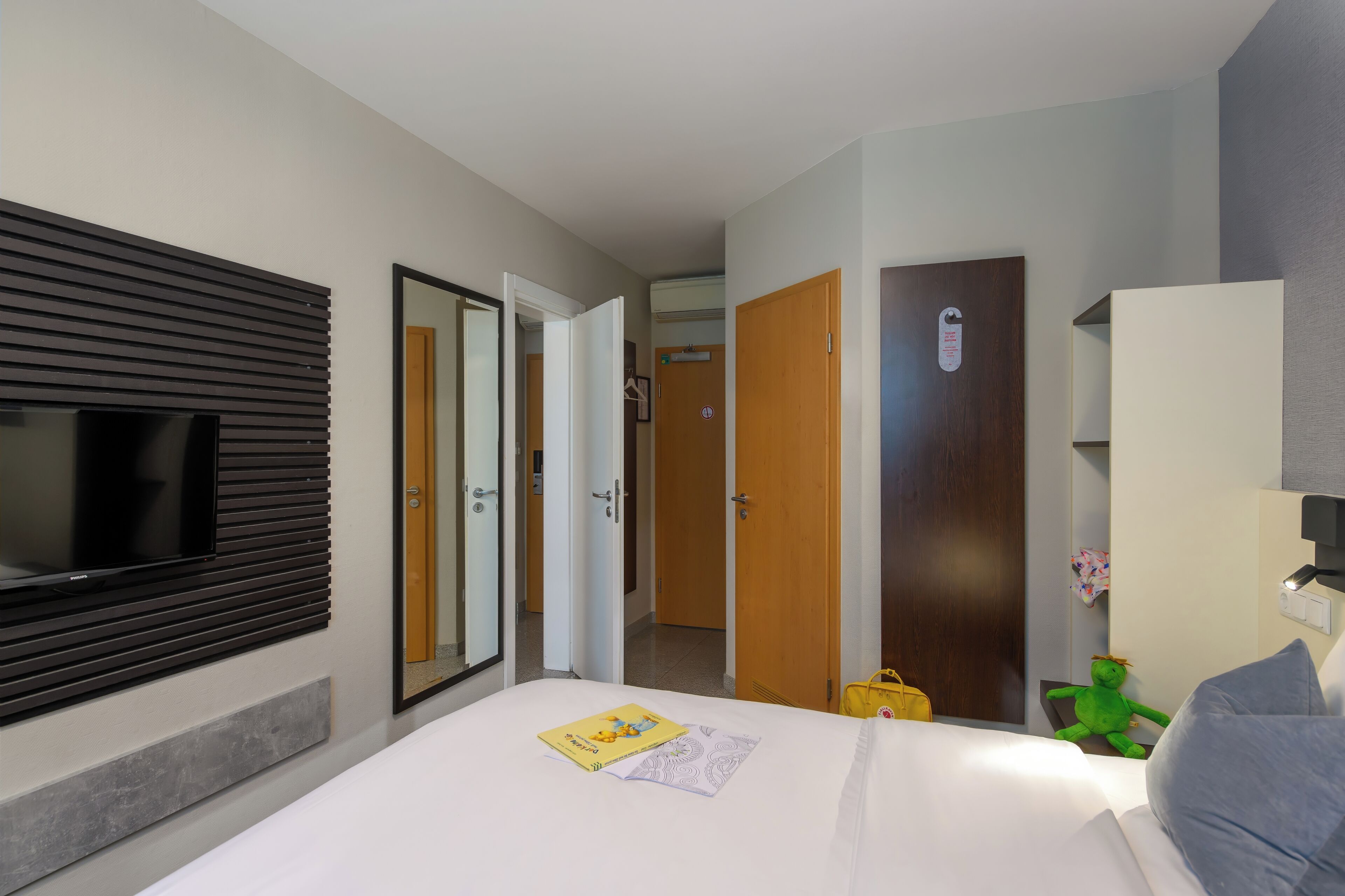 Une chambre d’hôtel avec un lit, un téléviseur fixé au mur, un miroir et une porte donnant sur une autre pièce.