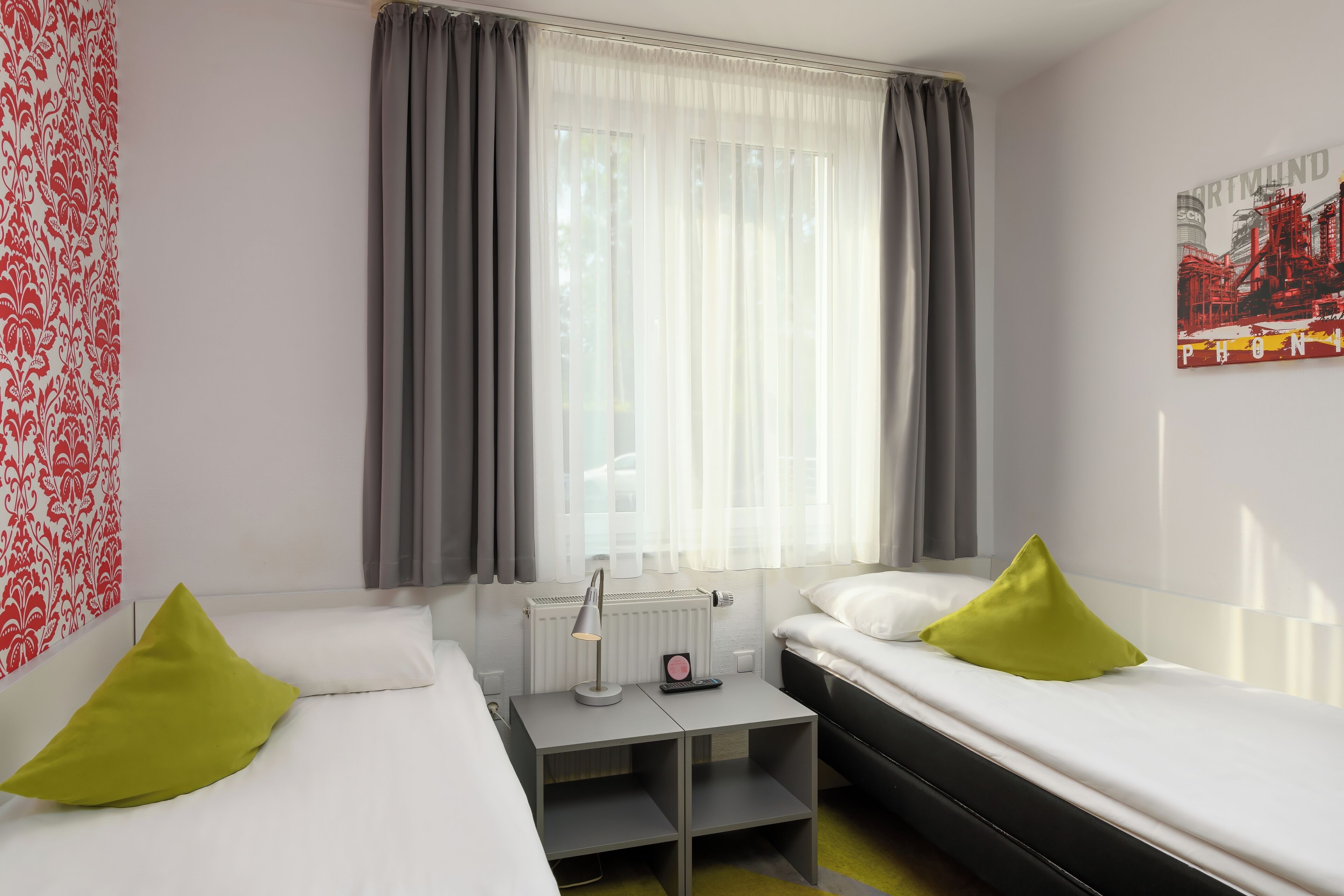 A hotel room with two beds, a small table with a lamp, a radiator, and a window with curtains.