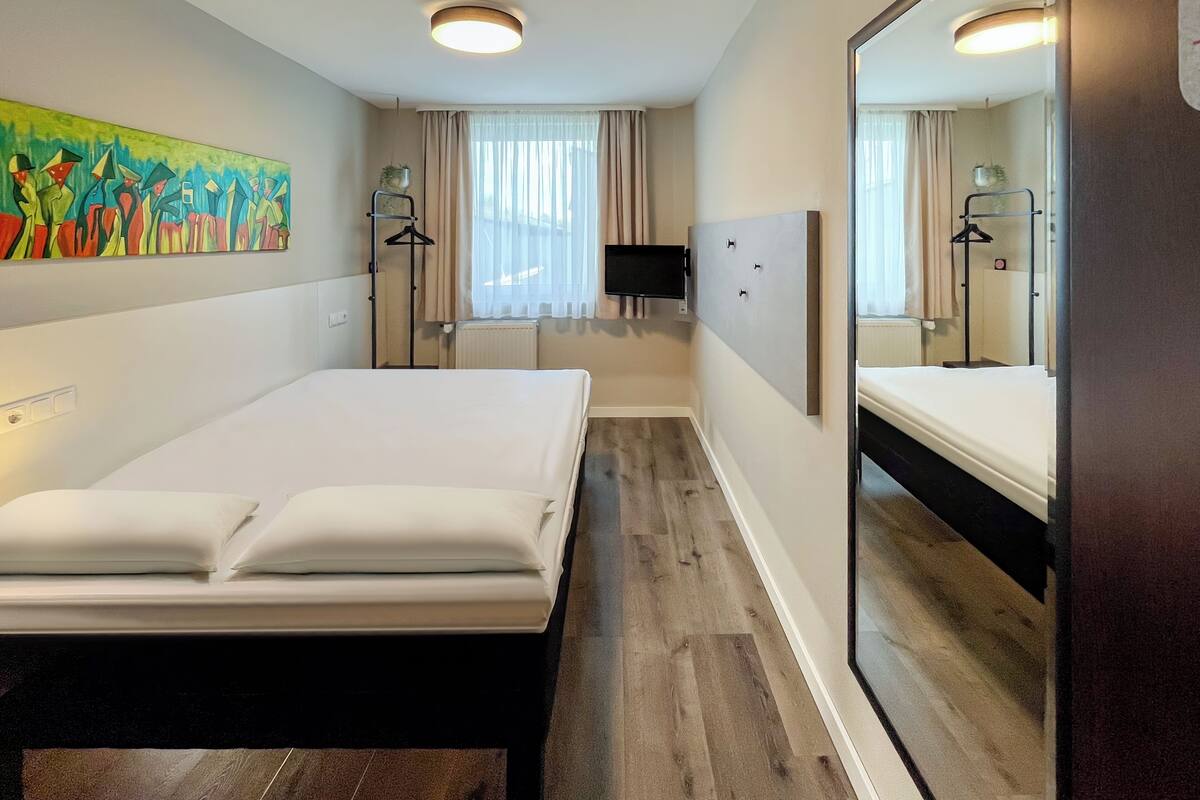 A hotel room with a large bed, a bedside table, a mirror, a painting on the wall, and a window with curtains.