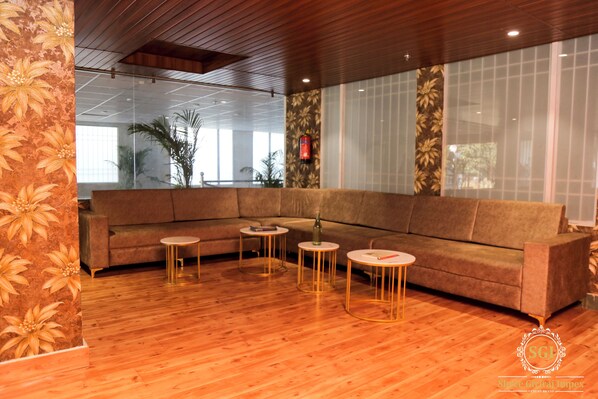A modern interior with a sectional sofa, three round tables, wooden flooring, and large windows with a view of palm trees. - Hotel SGI (Jaipur)