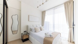 A modern bedroom with a large bed, a bedside table, a mirror, and a window with curtains. - Bright 1BR in Binghatti Corner (Dubai)