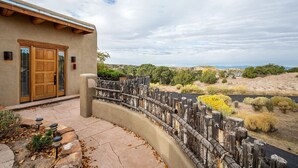 A stucco building with a wooden door and a curved stone pathway leading to a wooden fence overlooking a dry landscape with sparse vegetation.