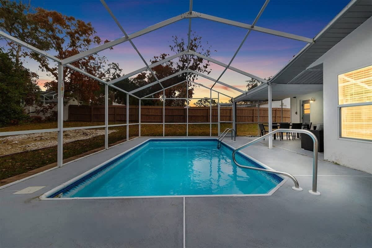 Ein überdachter Pool mit Metallrahmen, eine Terrassenfläche und Blick auf einen Wohngarten.