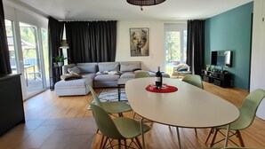 Una sala de estar moderna con un sofá, una mesa de comedor y sillas. Hay un cuadro en la pared, un televisor y una ventana con cortinas.