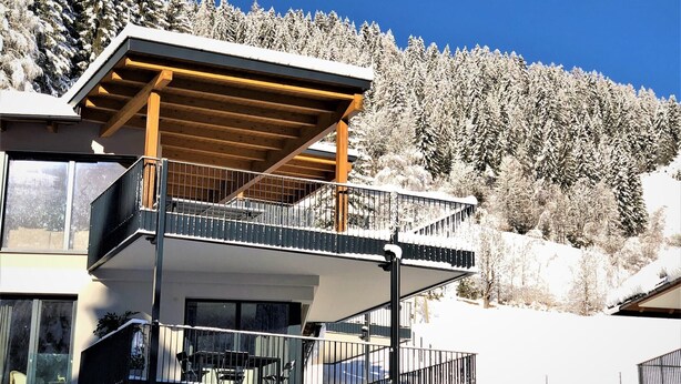 Un edificio de varios pisos con balcones, una zona exterior cubierta y un paisaje boscoso nevado al fondo.