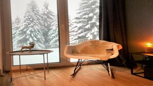 Una habitación con un gran ventanal, una mesa de madera con una pequeña figura de ciervo, una silla moderna y una vela encendida sobre una mesita auxiliar.