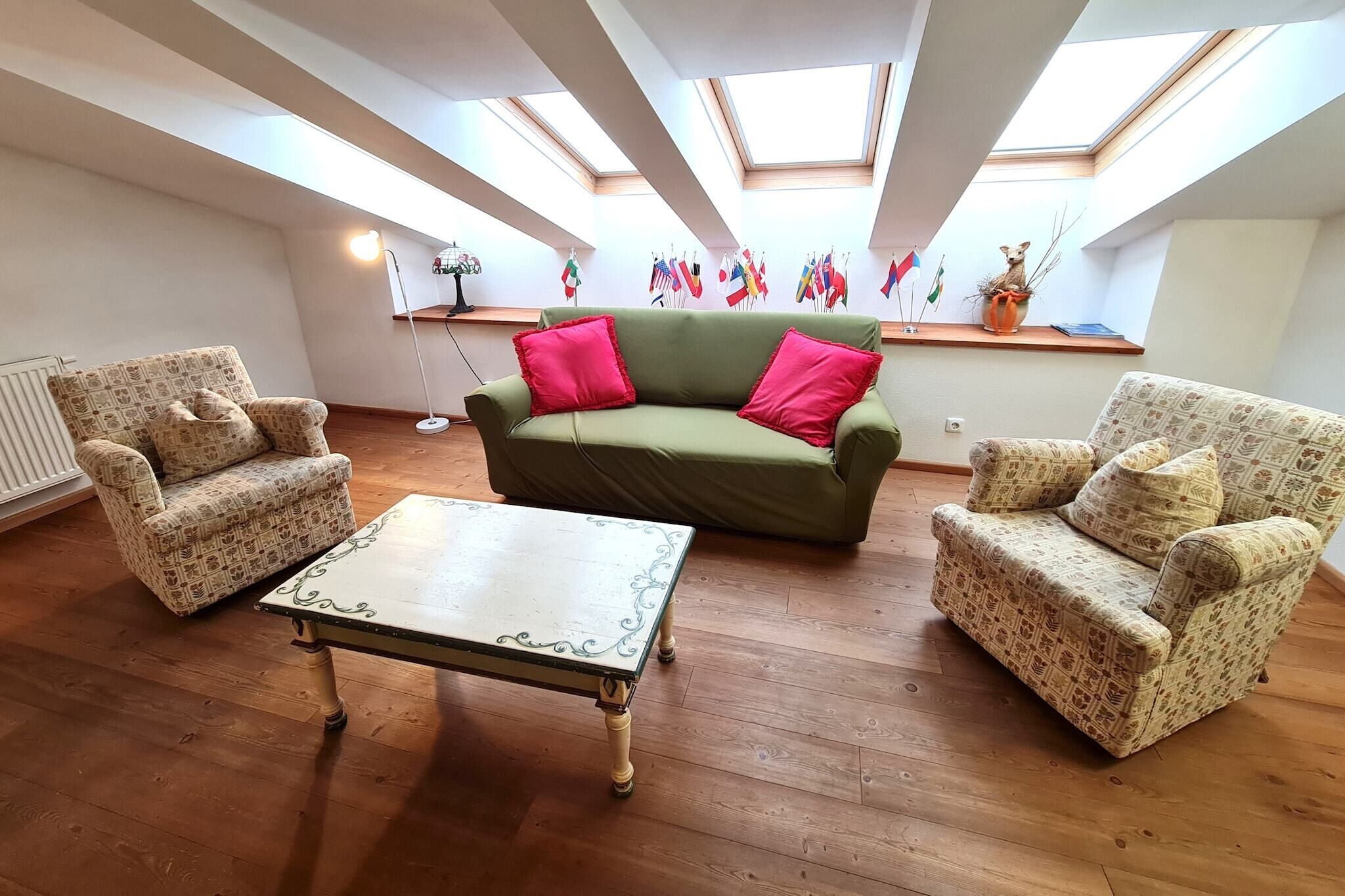 Uma sala de estar com sofá verde, poltronas estampadas e uma mesa branca. Há claraboias no teto, um radiador e um abajur. Bandeiras estão penduradas na parede.