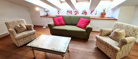 Uma sala de estar com sofá verde, poltronas estampadas e uma mesa branca. Há claraboias no teto, um radiador e um abajur. Bandeiras estão penduradas na parede.