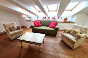 Un salón con un sofá verde, sillones estampados y una mesa blanca. Hay tragaluces en el techo, un radiador y una lámpara de pie. Se exhiben banderas en la pared.