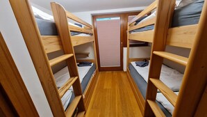 Een smalle kamer met stapelbedden langs de wanden, een houten vloer en een deur met een raam.