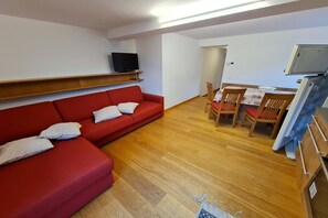 Una sala de estar con un sofá rojo, un televisor en una estantería, una mesa de comedor con sillas y un refrigerador.