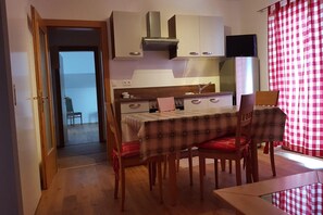 Une petite cuisine avec des meubles blancs, un fourneau et un téléviseur placé au-dessus du comptoir. On y trouve une table à manger avec quatre chaises, et un rideau à carreaux rouges et blancs à la fenêtre.