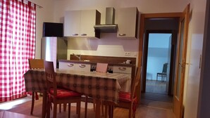 A compact kitchen with white cabinets, a stove, and a microwave. There is a dining table with a plaid tablecloth and red chairs. A TV is mounted above the stove.