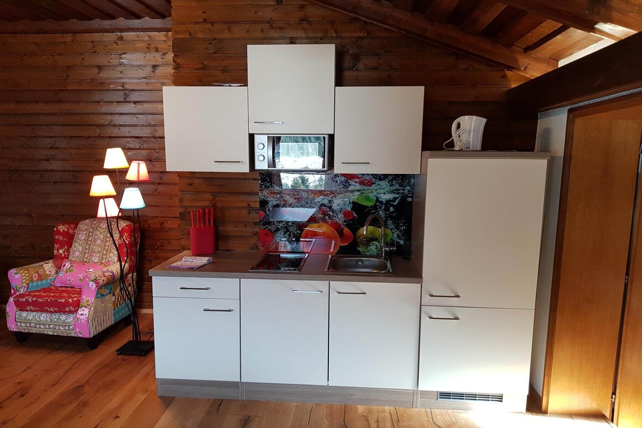 A compact kitchen with white cabinets, a microwave, and a refrigerator. There is a colorful chair with a floral pattern and a lamp with multiple shades.