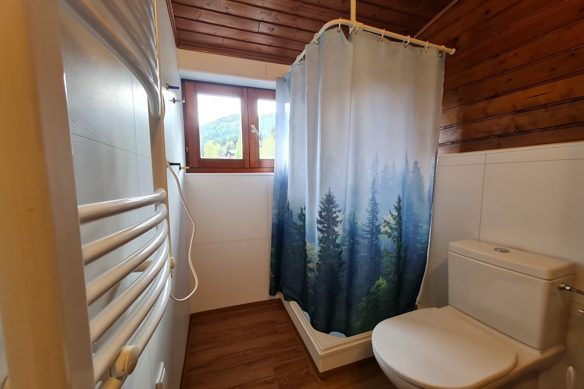 Une salle de bain avec un rideau de douche représentant une scène de forêt, des toilettes et une fenêtre donnant sur des arbres.