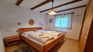 A wooden-framed bed with white linens and towels, a small wooden nightstand, a wall-mounted lamp, and a window with lace curtains. - Apartment with mountain views (Bad Kleinkirchheim)
