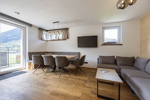En moderne stue med trægulv, en grå sofa, et spisebord med stole, et fjernsyn monteret på væggen og et stort vindue med udsigt til et bjerg.