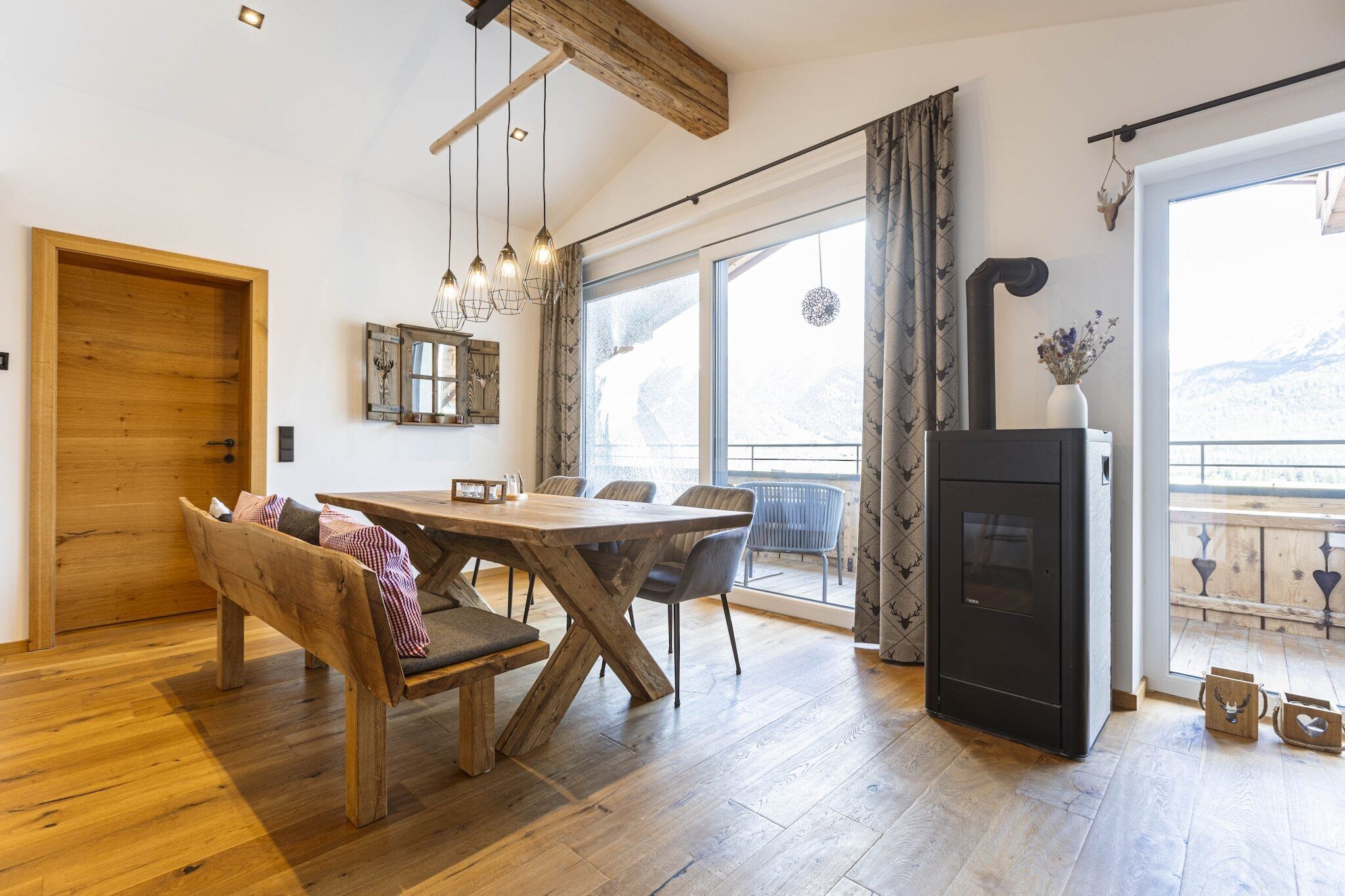 Una habitación acogedora con una mesa de comedor de madera, sillas y bancos. Hay una chimenea negra, un ventanal con cortinas y una puerta de madera.